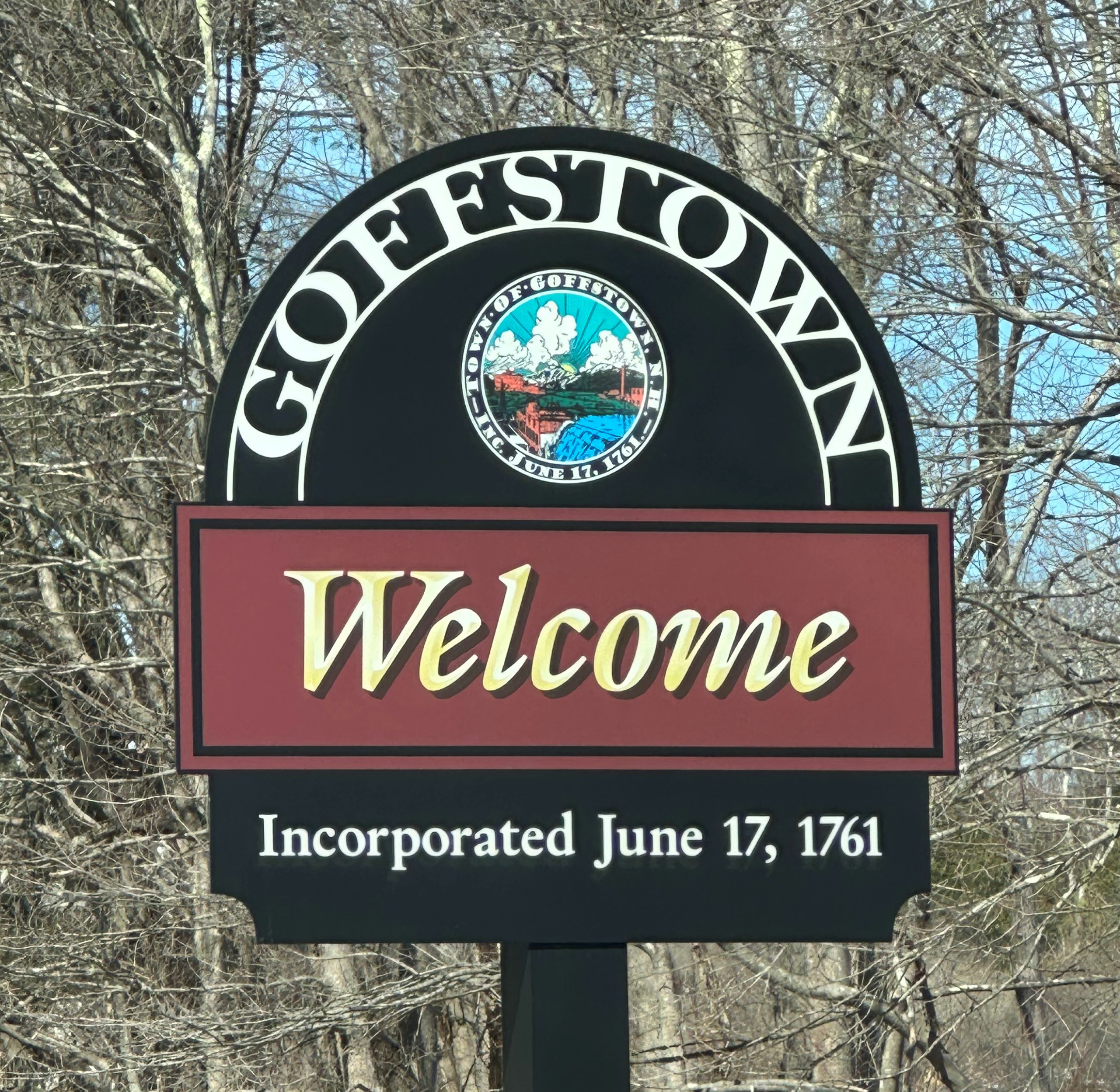 The photo of the Goffstown town sign is the word 'Goffstown' in an arch at the top of a large black sign, and at the base, it reads 'Incorporated June 17th, 1761.' Slightly above the bottom line is a large 'welcome' with a red background, and above that but below the Goffstown arch is a smaller circle with an illustrated picture of a small waterfall and sunrise, encircled by the words 'Town of Goffstown - Inc June 17, 1761'