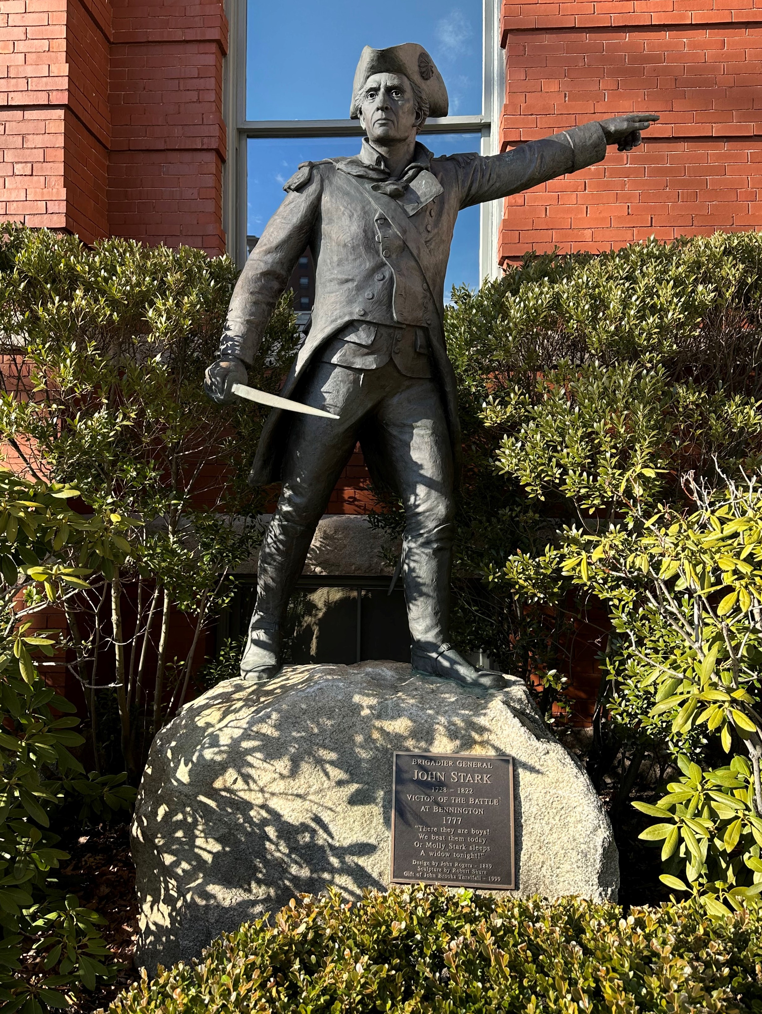 A photo of a statue of John Stark found in New Hampshire, the statue depicts him pointing to his left with his left hand raised above his neckline, and in his right hand he holds a short sword. He is dressed in a petticoat and hat. He is standing on a boulder, and on the boulder is a plaque that reads 'Brigadier General, John Stark, 1728-1822, Victor of the Battle at Bennington, 1777, 'There they are boys! We beat them today/ Or Molly, Stark sleep a widow tonight!' Design by John Rogers 1889, Sculpture by Robert Shure, Gift of John Brooks Threlfall 1999.'