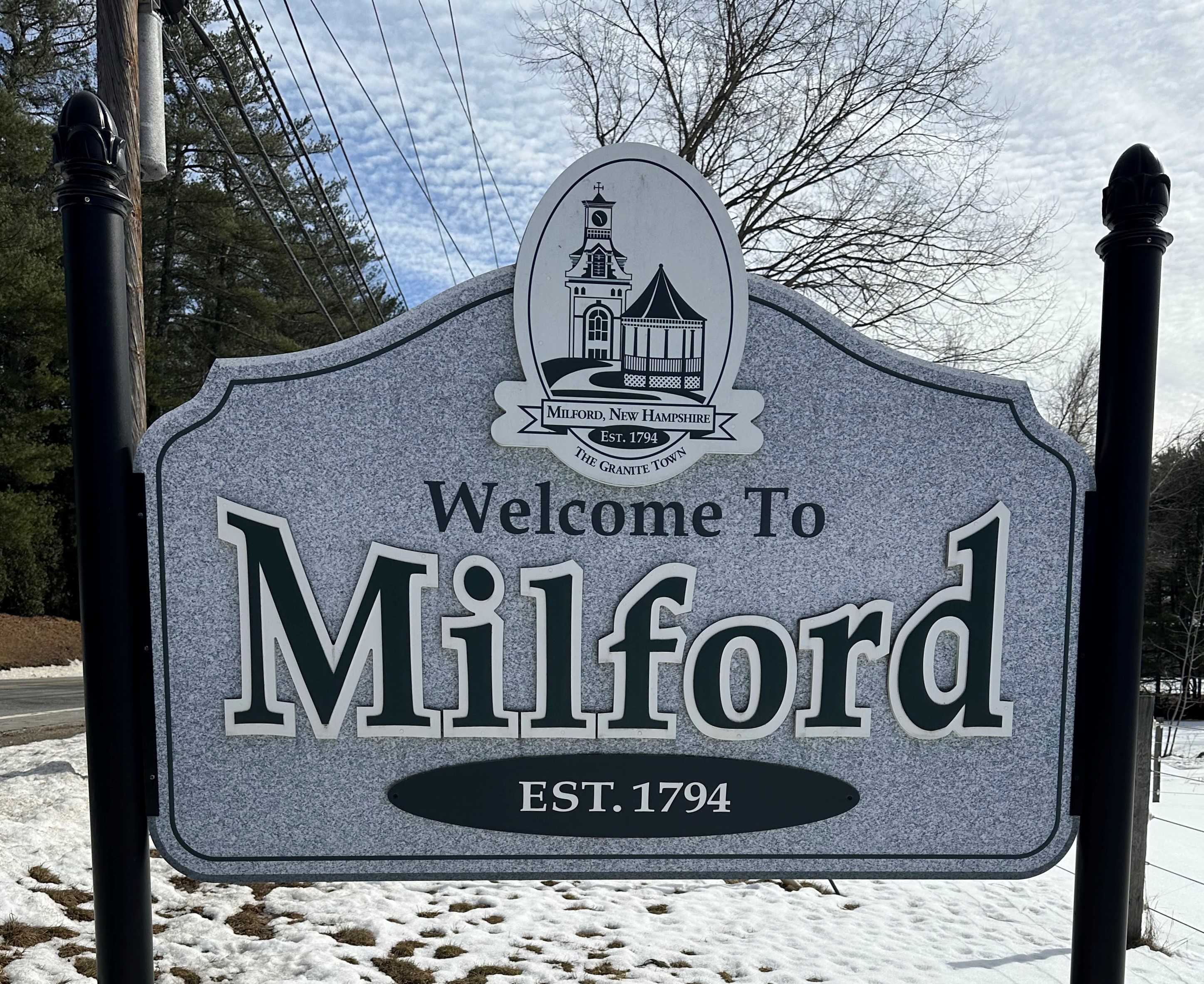 This is a photo of the Milford twon sign, which is held up by two black signposts, and which contains at the top an illustration of a gazebo and a church or similar towered building, a banner that says 'Milford, New Hampshire', and underneath that a small circle which reads 'est. 1794.' Underneath both of these is a small