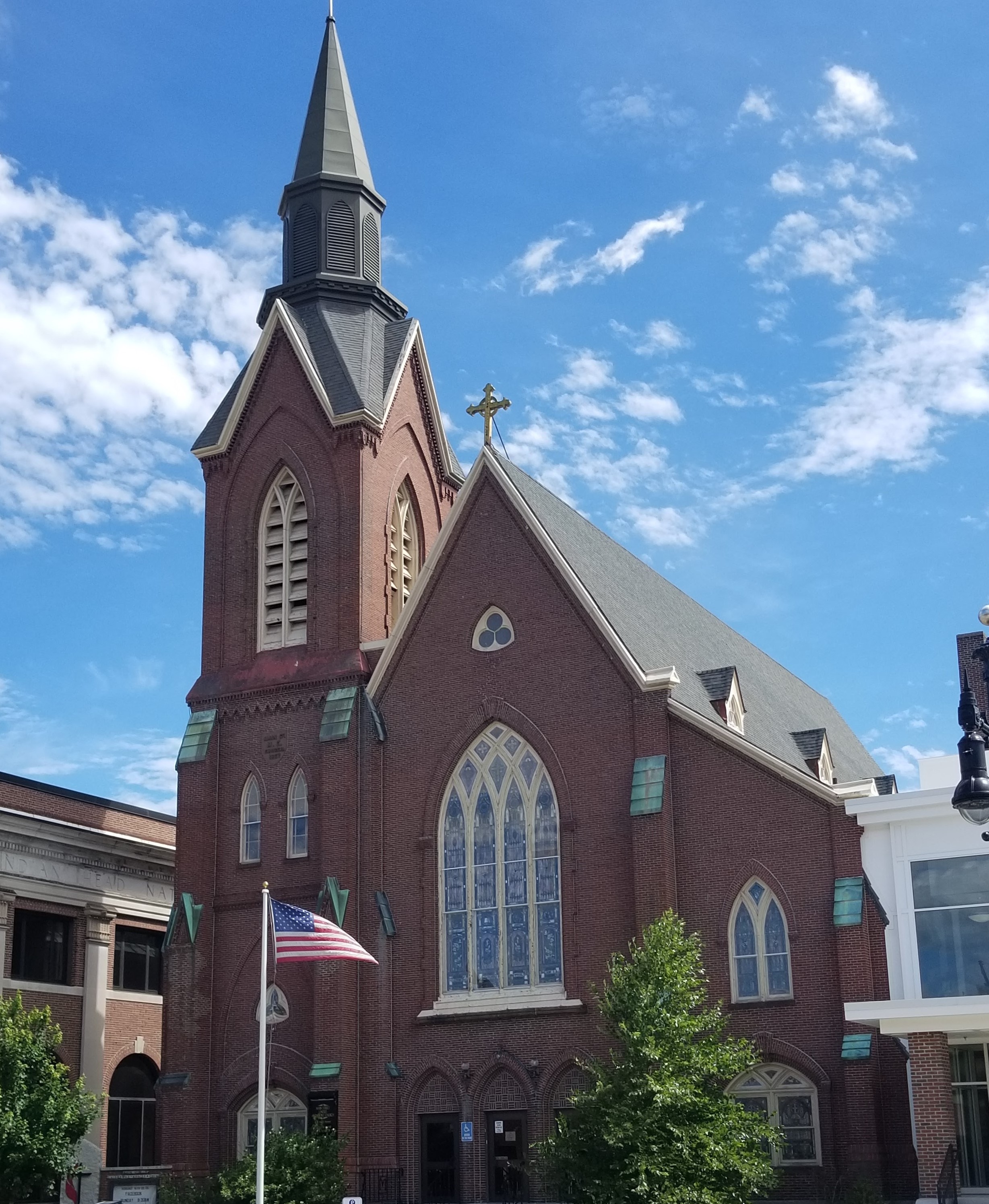 This is a photo of a Nashua, New Hampshire church. The church has a brick exterior, with one tall steeple and one main building. The windows have cream paneling and stained glass. The US flag is at full mast outside the church. The photo is taken from the ground perspective, so the mostly blue, slightly cloudy sky is visible behind the church. This photo is featured by New Hampshire personal injury law firm Buckley Law Offices.